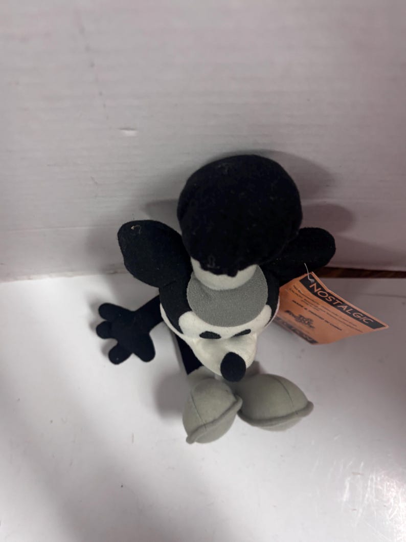 May include: A black and white plush toy mouse with large ears, a round head, and a white face. The mouse has black arms and a tail, with gray shoes. A tag that says "NOSTALGIC" is attached to the toy.
