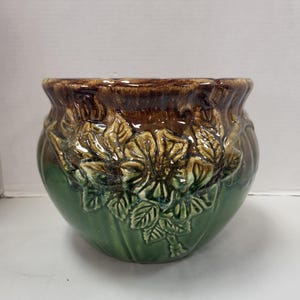 May include: A green and brown glazed ceramic planter with a floral design. The planter is round and has a wide opening.