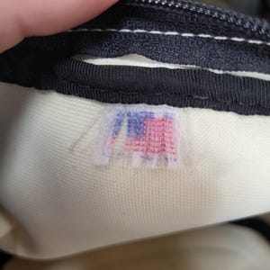 May include: Close-up of a black zipper and a cream-colored fabric. A small, faded tag with a red, white, and blue design is visible. The tag appears to be a logo or emblem.