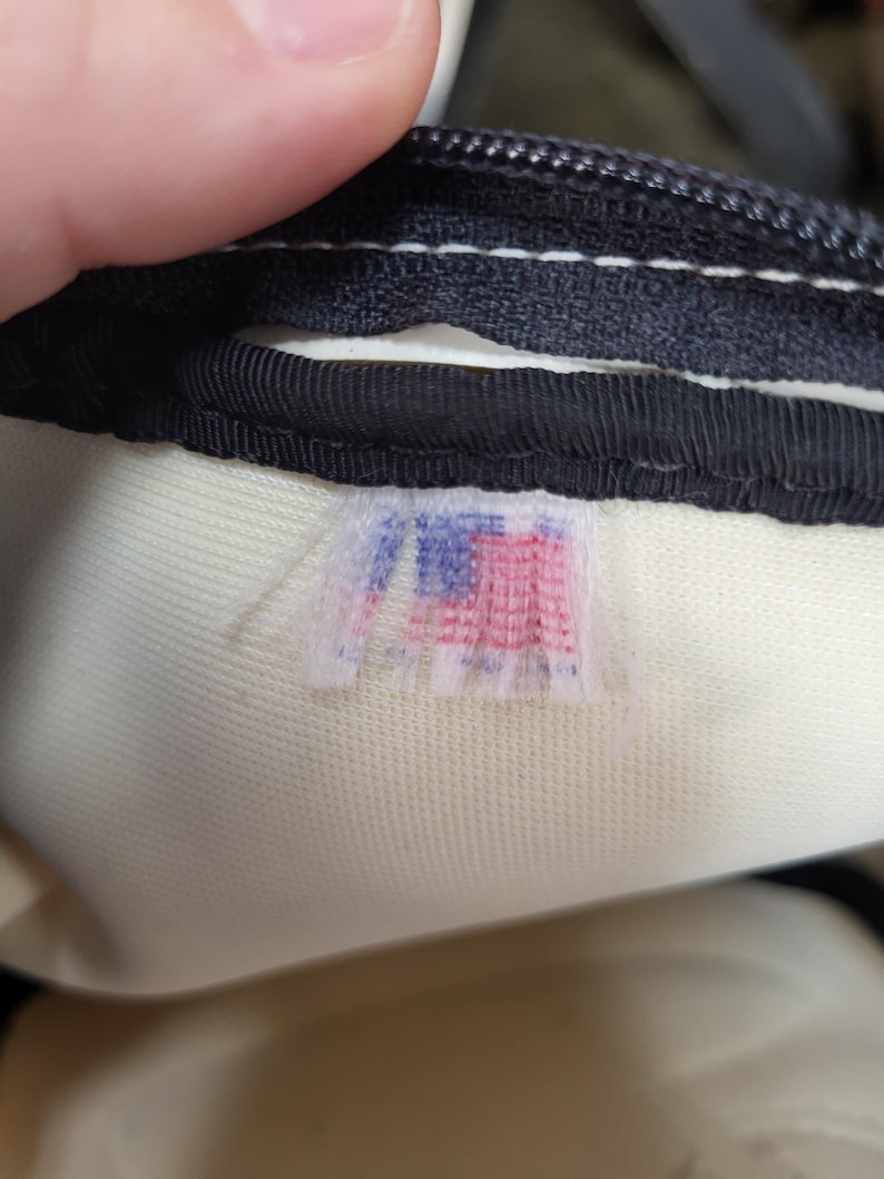 May include: Close-up of a black zipper and a cream-colored fabric. A small, faded tag with a red, white, and blue design is visible. The tag appears to be a logo or emblem.
