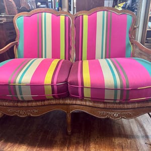May include: A two-seat sofa with a wooden frame and vibrant striped cushions. The cushions feature pink, green, yellow, and cream stripes. The sofa has curved armrests and decorative carvings on the base.