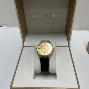 Pu&ograve; includere: Un orologio da polso Versace color oro con quadrante rotondo, finitura a raggiera e cinturino in pelle nera, presentato all'interno di una scatola bianca aperta con il marchio Versace stampato in lettere dorate sul coperchio interno.