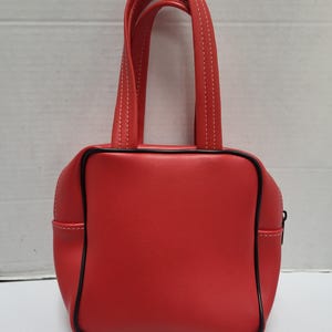 May include: A small, red handbag with a square shape and black trim. The bag has two short handles with white stitching and a zipper closure. The bag is made of a smooth material.