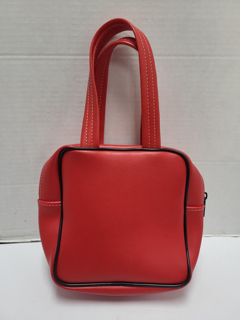 May include: A small, red handbag with a square shape and black trim. The bag has two short handles with white stitching and a zipper closure. The bag is made of a smooth material.