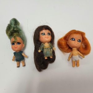 May include: Three small vintage dolls with painted faces and rooted hair. One doll has green hair and a dark blue dress, another has brown hair and a green dress, and the third has orange hair and white underwear. The dolls are on a white surface.