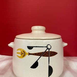 Peut inclure: Casserole en céramique blanche avec un couvercle, ornée d'un motif brun et noir d'ustensiles de cuisine, dont une fourchette, une cuillère et des cuillères à mesurer. Le motif est stylisé et abstrait.