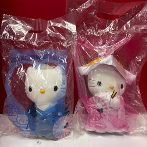 May include: Two Hello Kitty plush toys in clear plastic packaging. One wears a blue kimono, the other a pink kimono. Both have yellow noses and black eyes. The packaging has the word "Wedding" printed on it.