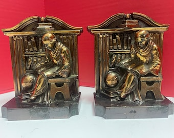 May include: Pair of bronze-toned bookends. Each bookend features a seated figure in a robe, reading, with a globe at their feet and a bookshelf behind them. The bookends have a rectangular base and are likely made of metal.