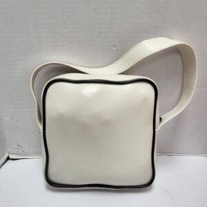May include: A square, off-white handbag with a black trim and a matching strap. The bag has a glossy finish and a simple, modern design. The strap is attached to the top of the bag.