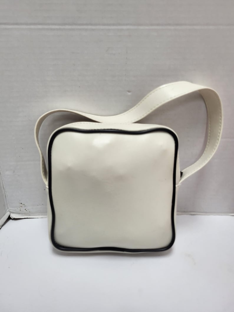 May include: A square, off-white handbag with a black trim and a matching strap. The bag has a glossy finish and a simple, modern design. The strap is attached to the top of the bag.