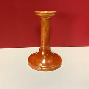 Puede incluir: Un portavelas alto de cerámica esmaltada en color naranja. Tiene una base ancha y redonda, un tallo delgado y cilíndrico, y una parte superior ensanchada. El color es un naranja cálido y brillante, con sutiles variaciones.