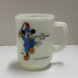 May include: White ceramic mug with a cartoon image of Mickey Mouse in a blue and white uniform playing a drum. The mug has the text "The Mickey Mouse Club" and "1955" printed on it.