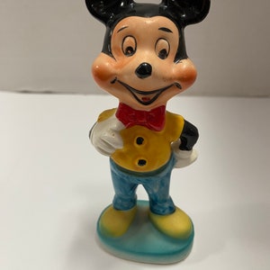 May include: A vintage ceramic figurine of Mickey Mouse wearing a yellow shirt, blue pants, and a red bow tie. He is standing on a blue base.
