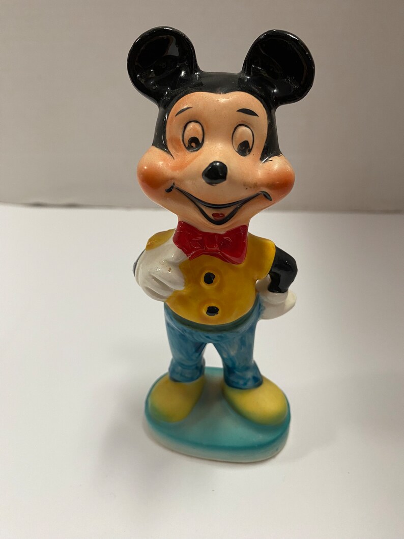 May include: A vintage ceramic figurine of Mickey Mouse wearing a yellow shirt, blue pants, and a red bow tie. He is standing on a blue base.