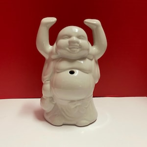 May include: White ceramic figurine of a smiling Buddha with raised arms. The figurine has a hole in the top of its head.