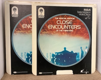 May include: Two white cardboard cases for a RCA SelectaVision VideoDisc. The front of the cases feature a black and white image of a crowd of people silhouetted against a red and orange background. The text on the case reads "The Special Edition Close Encounters of the Third Kind".