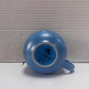 May include: A light blue ceramic mug with a handle. The bottom of the mug features the Disney logo and text. The mug is resting on a white surface.