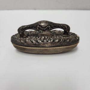 May include: An oval-shaped silver-toned clothes brush featuring an ornate, embossed floral pattern on the handle and top. The handle is arched and integrated into the decorative metal top, which sits above a light-colored, textured bristle base.