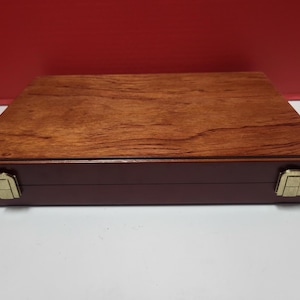 Puede incluir: Una caja rectangular de madera con un acabado marrón rojizo y una parte superior de veta de madera más clara. La caja tiene una base marrón oscuro y cierres dorados. La caja está cerrada y se encuentra sobre una superficie blanca.