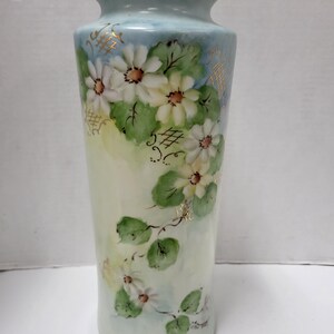 May include: A tall, hand-painted vase with a light blue background and a gold rim. The vase features white daisies with yellow centers and green leaves. Delicate gold accents and floral details adorn the vase, creating an elegant, vintage aesthetic.