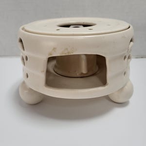 May include: Off-white ceramic tea warmer with a circular design. The warmer has a round top with several small holes and a central opening. It features a cylindrical body with cut-out details and rests on three spherical feet.