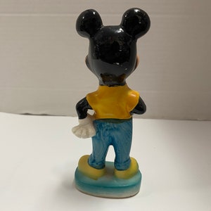 May include: A ceramic figurine of Mickey Mouse wearing a yellow shirt and blue jeans. He is standing on a blue base.