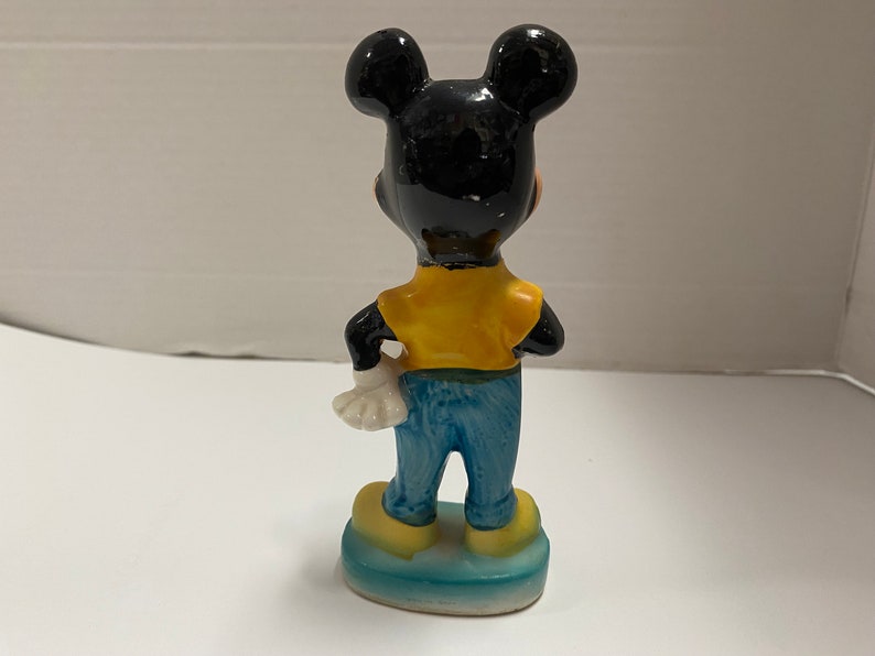 May include: A ceramic figurine of Mickey Mouse wearing a yellow shirt and blue jeans. He is standing on a blue base.