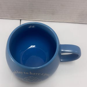 May include: A blue ceramic mug with a rounded shape and a handle. The mug has the phrase "good day to have a good" printed in white script near the base. The interior of the mug is a darker shade of blue.