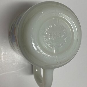 May include: White ceramic mug with a blue and orange floral design. The bottom of the mug has the text "Fire King Ovenware Made in USA" printed on it.