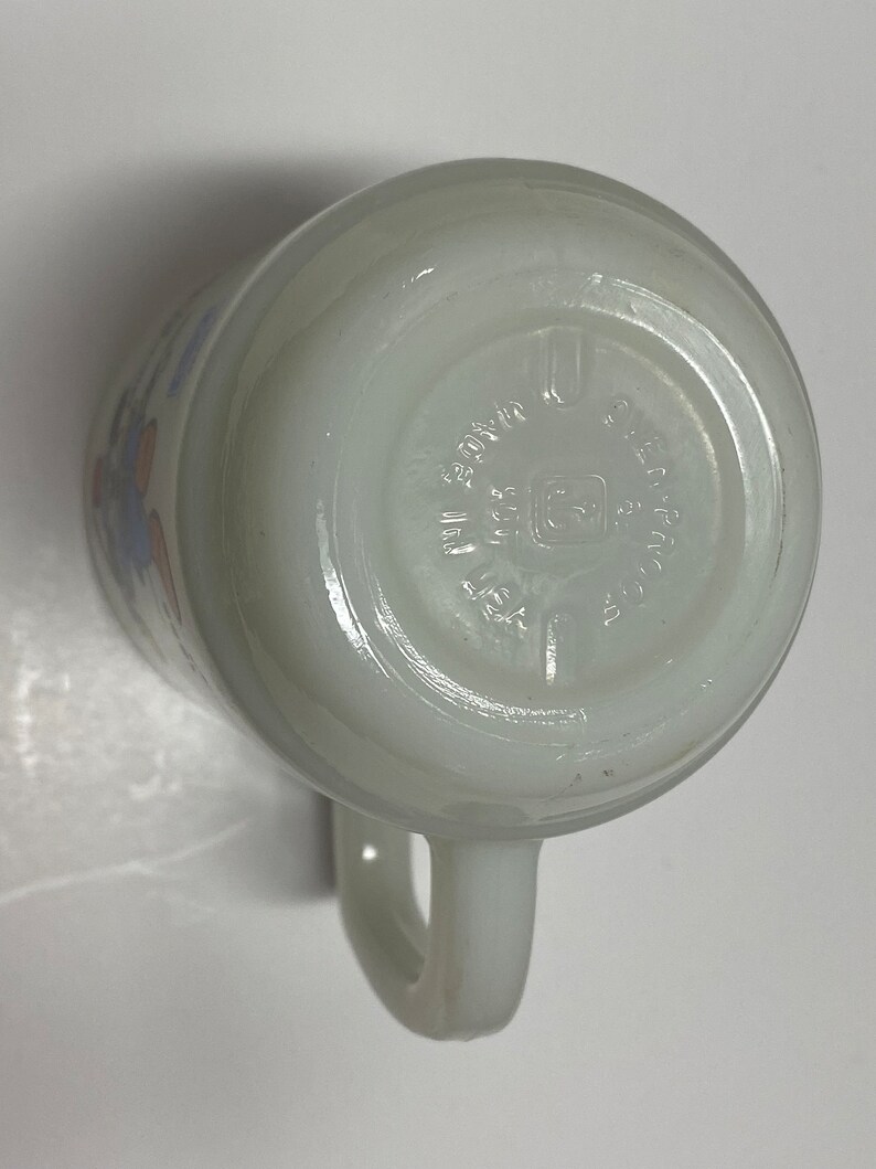 May include: White ceramic mug with a blue and orange floral design. The bottom of the mug has the text "Fire King Ovenware Made in USA" printed on it.