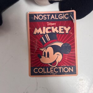 May include: A rectangular tag with the words "NOSTALGIC" and "MICKEY" in red and gold lettering. The tag features a vintage illustration of Mickey Mouse wearing a top hat. The word "COLLECTION" is at the bottom.