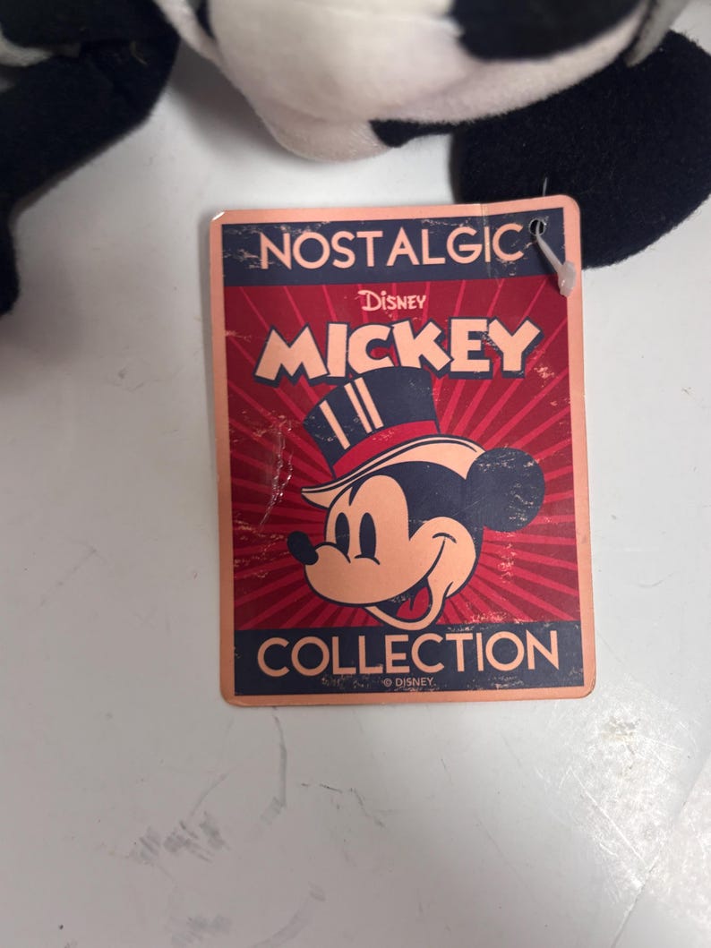 May include: A rectangular tag with the words "NOSTALGIC" and "MICKEY" in red and gold lettering. The tag features a vintage illustration of Mickey Mouse wearing a top hat. The word "COLLECTION" is at the bottom.