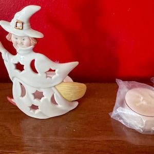 Könnte beinhalten: Weiße Keramik-Halloween-Hexenfigur, die einen Besen hält, mit einem gelben Besen und einem weißen Teelichthalter. Die Hexe trägt einen weißen Hut mit einer goldenen Schnalle. Der Teelichthalter befindet sich in einer durchsichtigen Plastikverpackung.