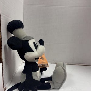 May include: A plush Mickey Mouse toy in black, white, and gray, seated with legs extended. The toy has a chef's hat and a tag that reads "The Disney Store Collection."