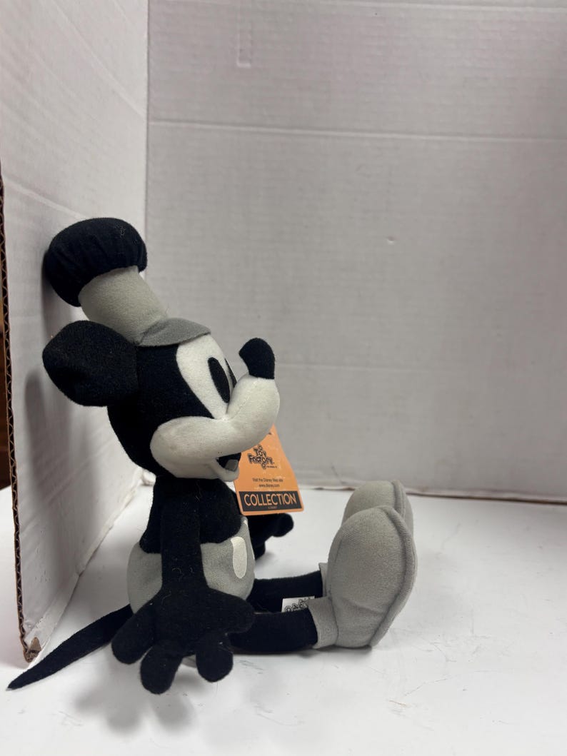 May include: A plush Mickey Mouse toy in black, white, and gray, seated with legs extended. The toy has a chef's hat and a tag that reads "The Disney Store Collection."