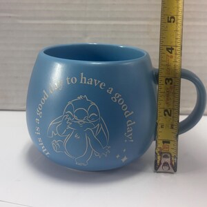 May include: A light blue ceramic mug with a handle. The mug features a white illustration of a cartoon character and the text "this is a good day to have a good day!". The mug is approximately 3 inches tall.