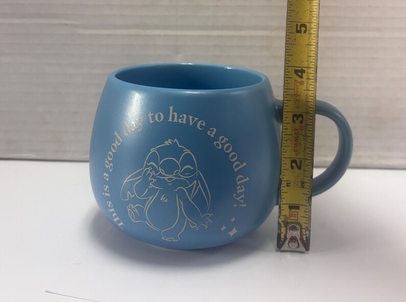 May include: A light blue ceramic mug with a handle. The mug features a white illustration of a cartoon character and the text "this is a good day to have a good day!". The mug is approximately 3 inches tall.