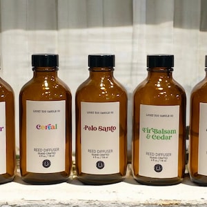 May include: Five amber glass bottles with black lids and white labels. The labels have the text "LOCKY YOU CANDLE CO" and the names of different scents: "Lavender Matcha", "Cereal", "Palo Santo", "Fir Balsam & Cedar", and "Smoke & Ember". Each bottle is labelled "Reed Diffuser" and "Island Crafted".