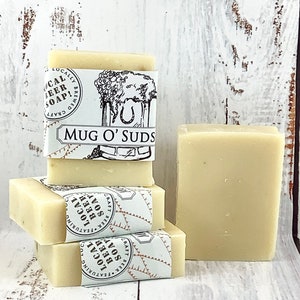 May include: Four bars of white soap stacked on top of each other. Each bar is wrapped in a white label with black text that reads "Craft Beer Featuring Local Beer Soap!" and "Mug O' Suds".