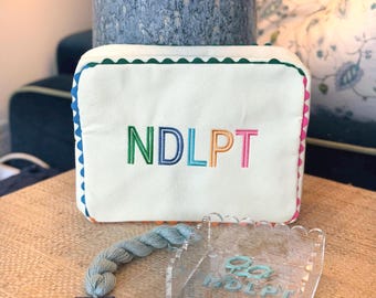 May include: A white rectangular pouch with the embroidered letters "NDLPT" in green, blue, orange, and pink. The pouch has a green scalloped edge and is trimmed with blue and pink scalloped borders. A clear acrylic organizer and a skein of blue thread are also present.