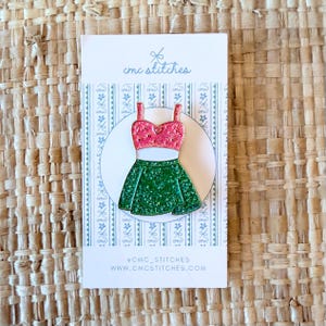 May include: A decorative pin featuring a pink and green dress design. The dress has a pink top and a green skirt. The pin is on a white card with the text "cmc stitches" and the website address.
