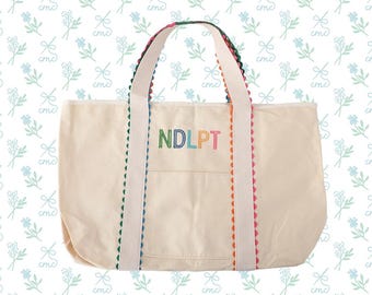 May include: Cream-colored tote bag with white handles and colorful trim. The bag features a front pocket and the embroidered letters "NDLPT" in rainbow colors. The background has a repeating pattern of blue flowers and bows.