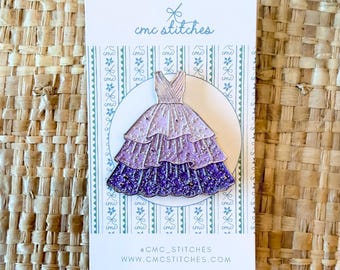 May include: A pin featuring a tiered, purple and lavender dress with glitter accents. The pin is on a white card with the text "cmc stitches" and social media information. The background has a repeating pattern of bows.
