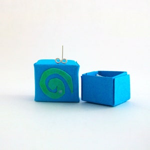 Op de afbeelding: Een klein blauw doosje met een groen swirl-ontwerp aan de voorkant. Het doosje is open en er zit een zilveren oneindigheidssymbool oorsteker in.