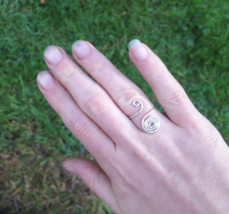 May include: A silver wire ring with a spiral design. The ring is worn on the middle finger of a hand.