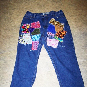May include: Blue denim jeans with a patchwork design on the front pockets. The patches are made of various fabrics and colours, including pink, red, blue, green, and yellow.