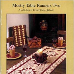 Pode incluir: Um livro intitulado "Mostly Table Runners Two: A Collection of Twenty Classic Patterns" com uma foto de um corredor de mesa vermelho e branco em uma mesa de madeira. O corredor de mesa é feito de tecido com um padrão geométrico.