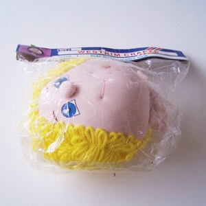 Könnte beinhalten: Ein Puppenkopf mit einem rosa Gesicht und blauen Augen. Die Puppe hat gelbes Garnhaar und ist in Plastik verpackt.
