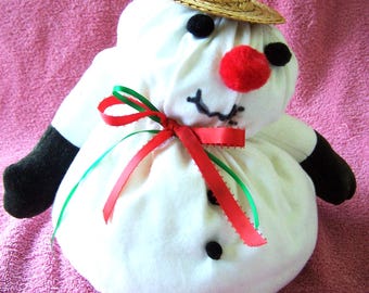Könnte beinhalten: Ein weißer Schneemann mit roter Nase und schwarzen Augen, der einen Strohhut und ein rot-grünes Band trägt. Der Schneemann sitzt auf einem rosa Handtuch.
