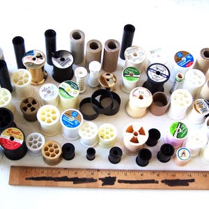 May include: A collection of sewing spools and bobbins in various sizes and colors, including black, white, beige, and gold. A wooden ruler is at the bottom. The spools are filled with thread, and some have labels.